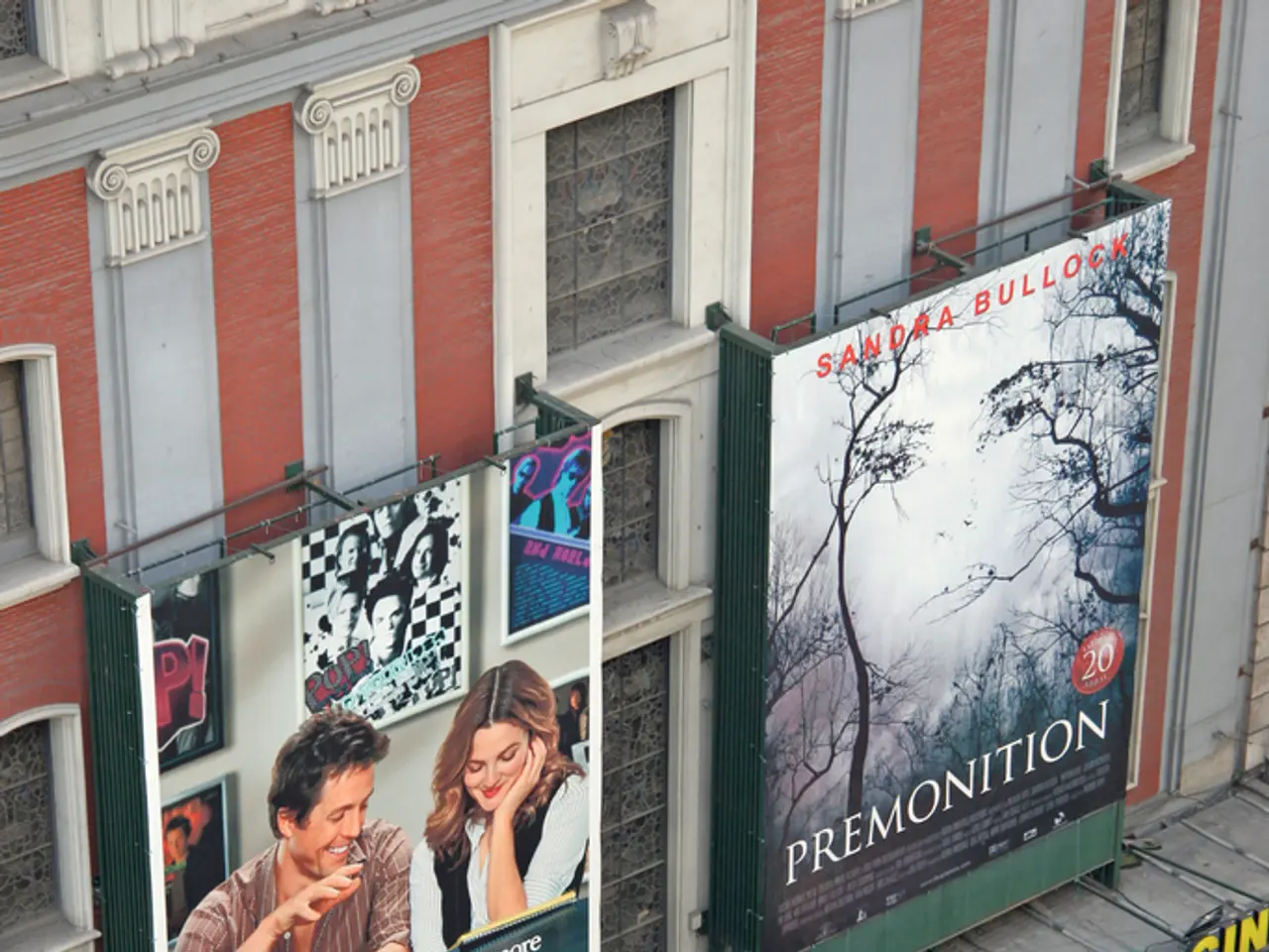 In this image there is a building in the middle. To the building there are two hoardings. At the...