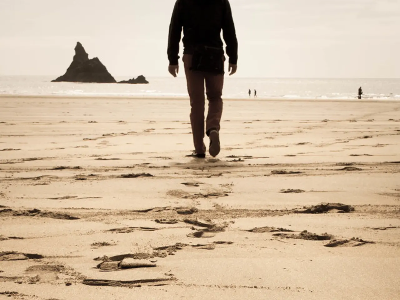 In the image there is a person walking on beach, in the front there is a small hill in the ocean...