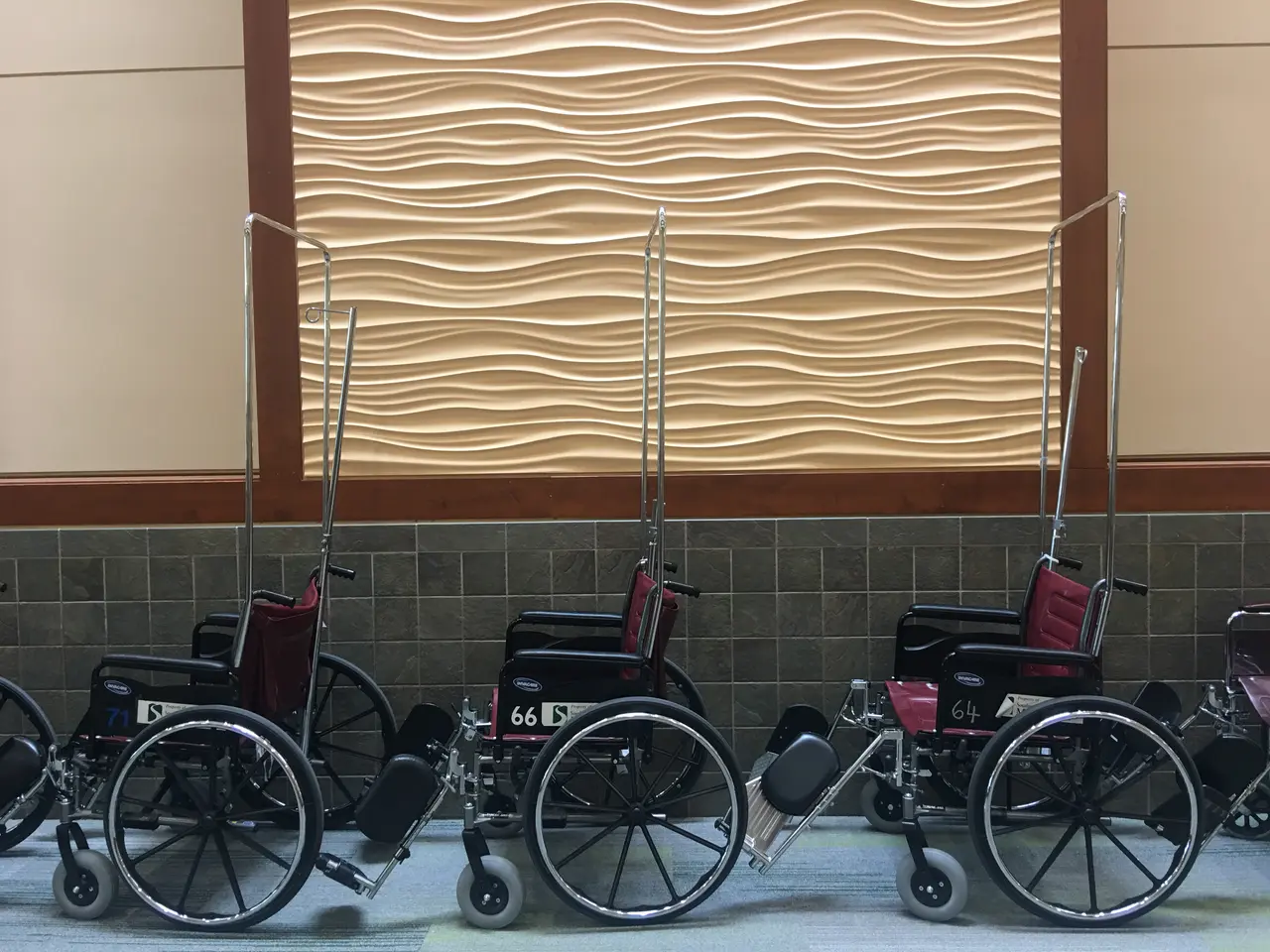 The image shows a row of wheelchairs lined up in a row in front of a wall with a design on it. The...