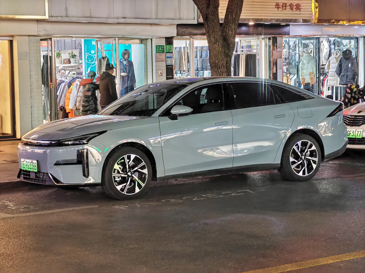The image shows an electric car parked on the side of a street at night, surrounded by buildings,...