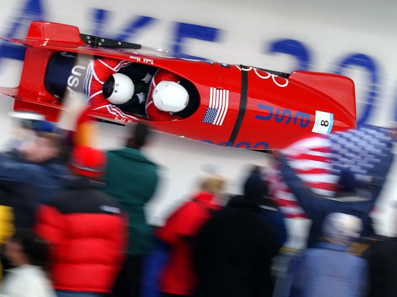The image shows a bobsled in action at the Olympics, with two people wearing helmets in the vehicle...