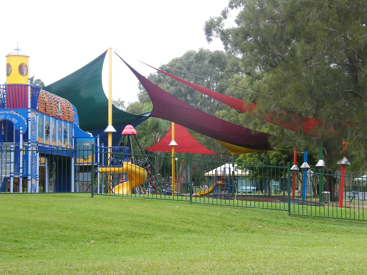 The image shows a park with a playground in the middle of it, surrounded by a metal fence and lush...