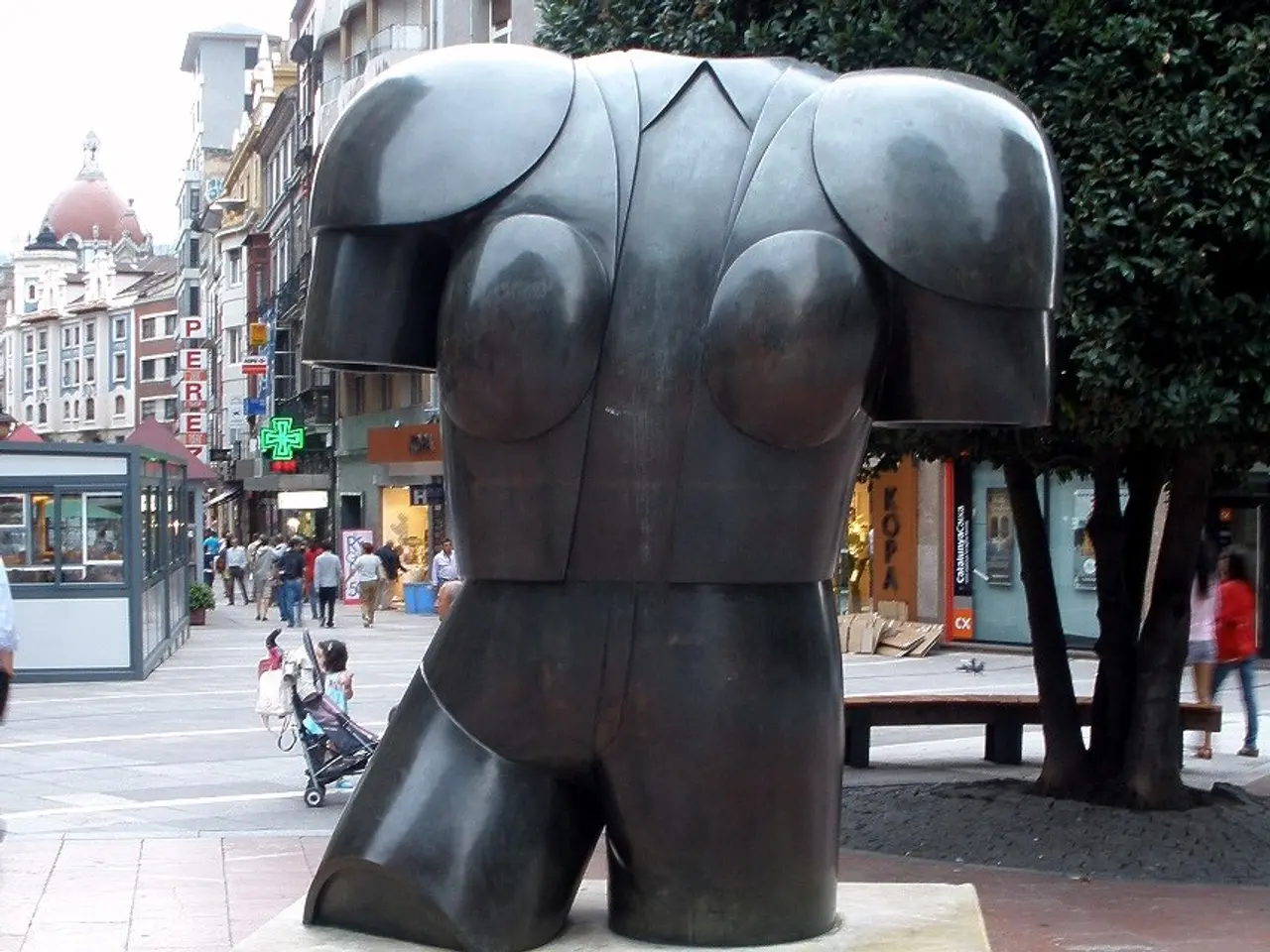 The image shows a large bronze sculpture of a woman's torso on a pedestal in the middle of a city...