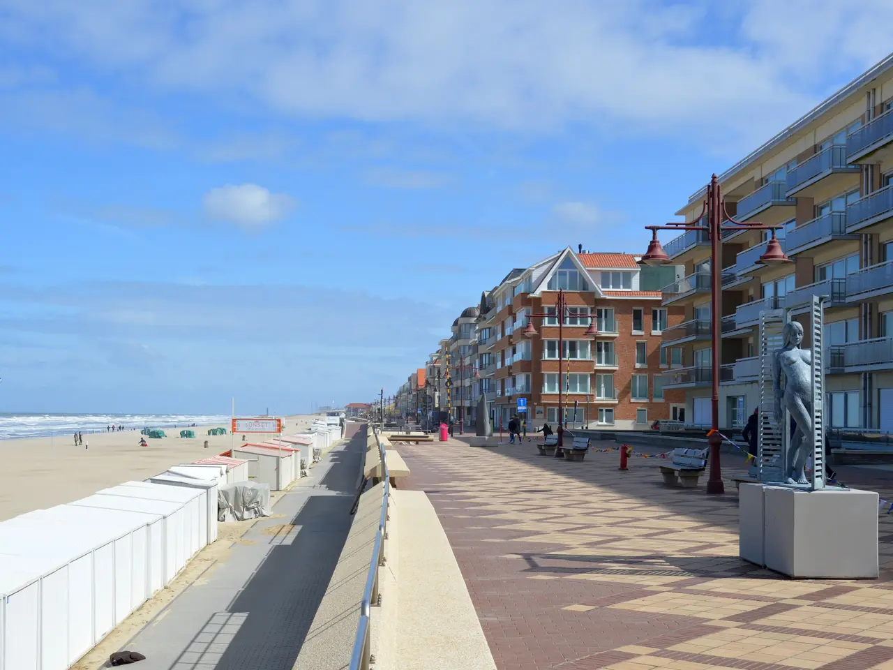 The image shows a promenade along the beach with people walking along it. On the right side of the...