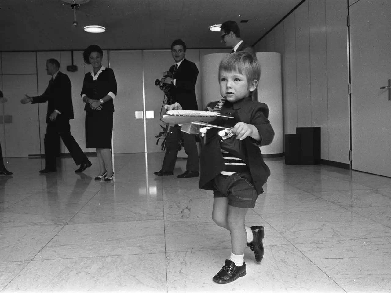 The image shows a young boy running through an airport with a toy airplane in his hand. He is...