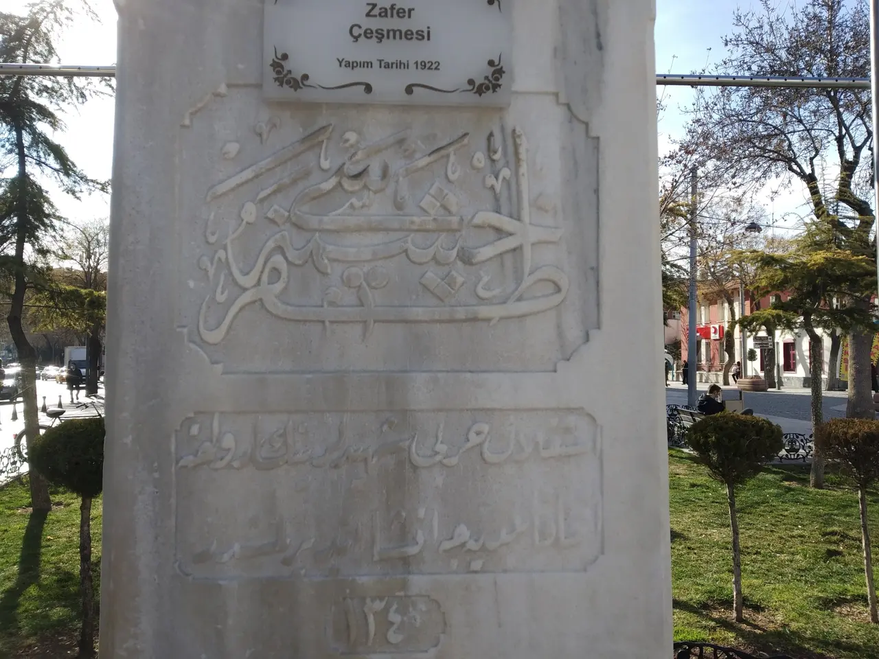 The image shows a monument in the middle of a park with Arabic writing on it. Surrounding the...