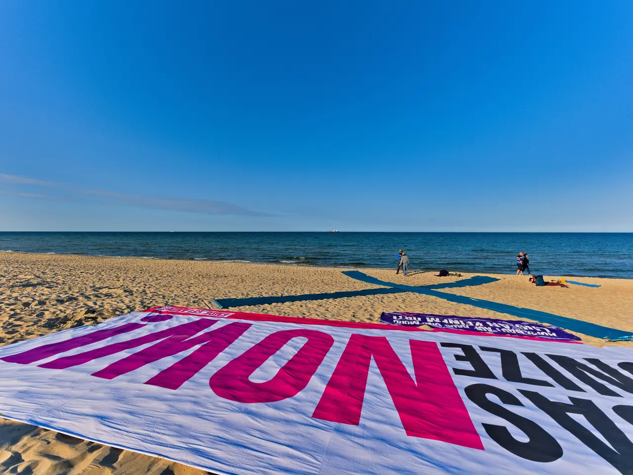 The image shows a banner on the beach with people walking on it, surrounded by sand and the vast...