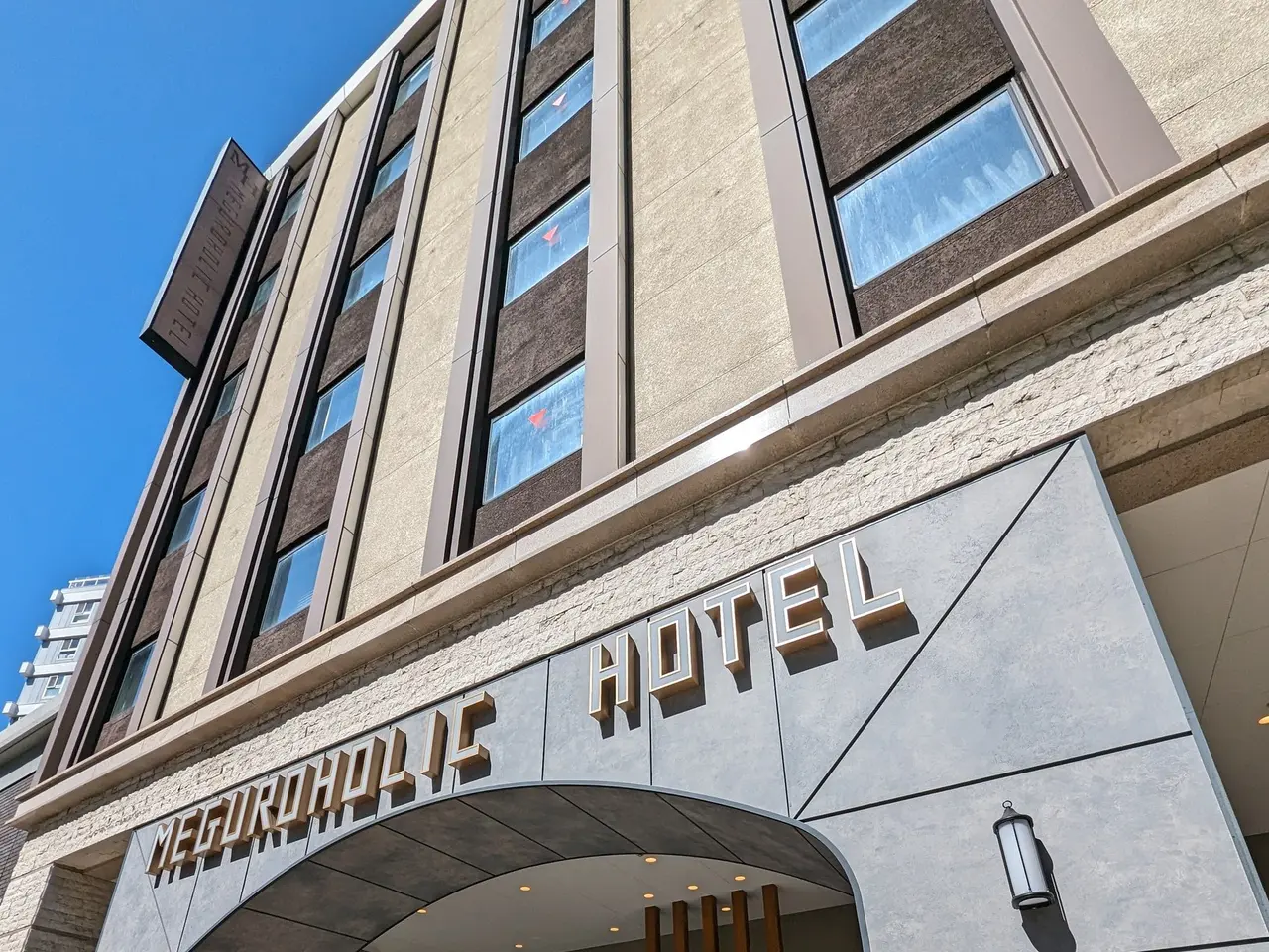 The image shows the entrance to the Meguroholic Hotel in Tokyo, Japan. It is a large building with...