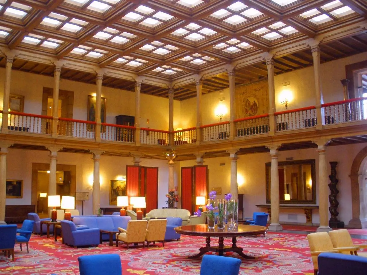 The image shows a large room filled with lots of blue chairs, sofas, tables, flower vases, lamps,...