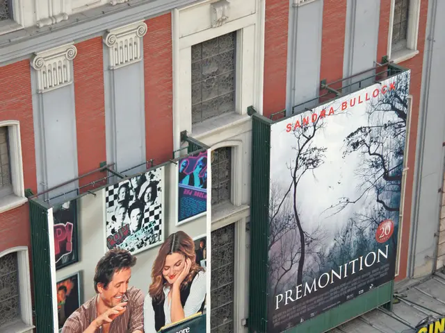 In this image there is a building in the middle. To the building there are two hoardings. At the...