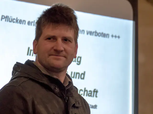 In this image there is a man standing. He is smiling. Behind him there is a projector board. There...