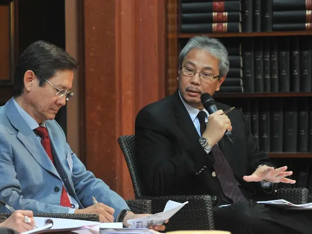 In this image there are three people sitting on chairs, one is holding a mike in his hand and...