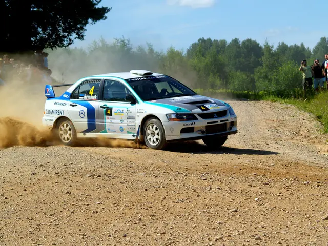 The image shows a rally car driving down a dirt road surrounded by a crowd of people, some of whom...