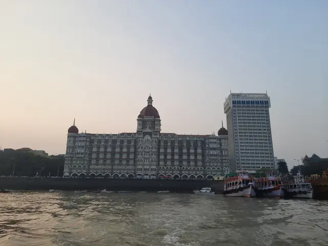 The image shows the Taj Mahal Palace Hotel in Mumbai, India, with a few boats on the water in front...