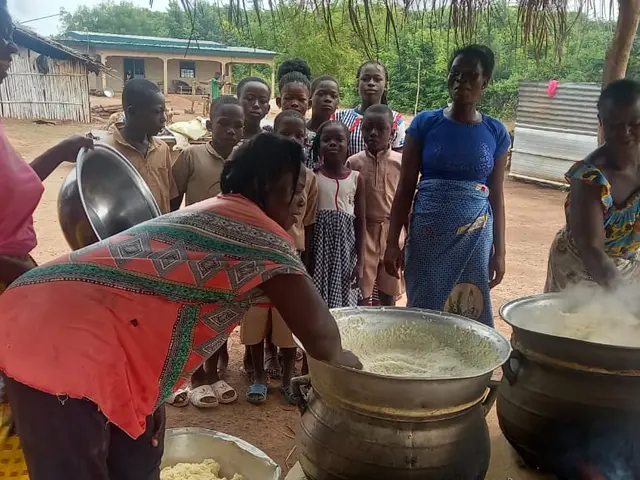 The image shows a group of people standing around a large pot filled with food, with a woman...
