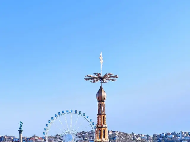 The image shows a bustling city square with a ferris wheel in the middle, surrounded by buildings,...