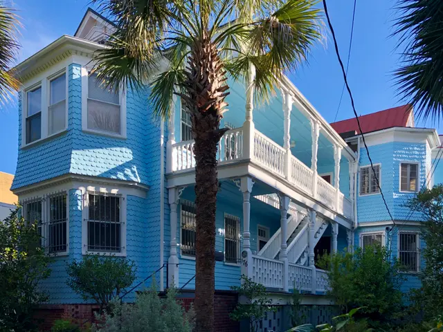 The image shows a blue house with a palm tree in front of it, surrounded by lush green plants and...