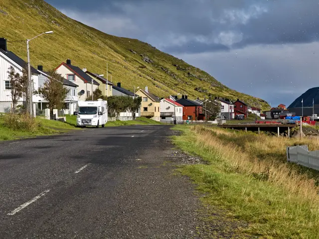 The image shows a truck driving down a road next to a small town in Iceland. On either side of the...