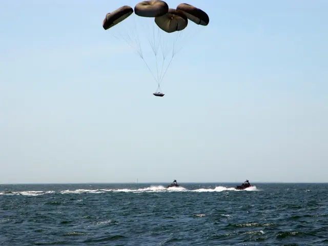 The image shows two people parasailing in the ocean with a boat in the background. The sky is...