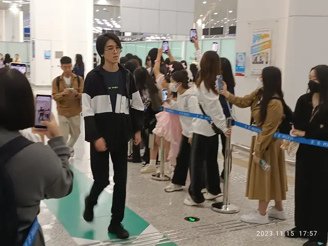 The image shows a group of people standing in line at an airport, some of them holding mobile...