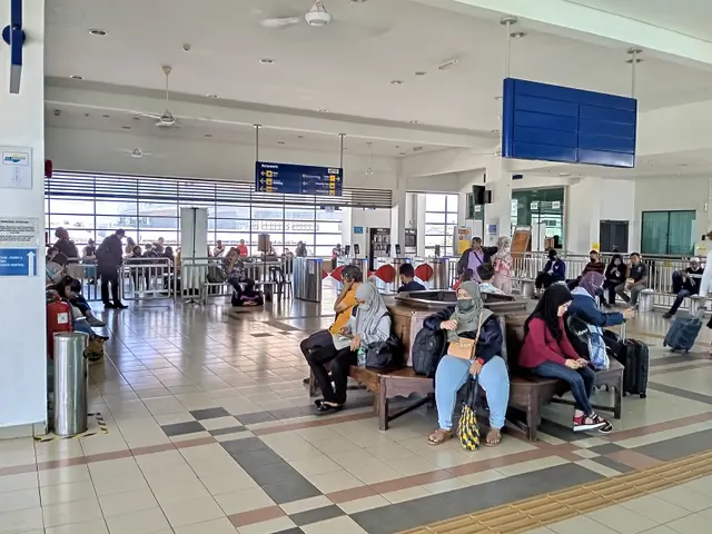 The image shows a group of people sitting on benches in an airport, with some of them holding bags....