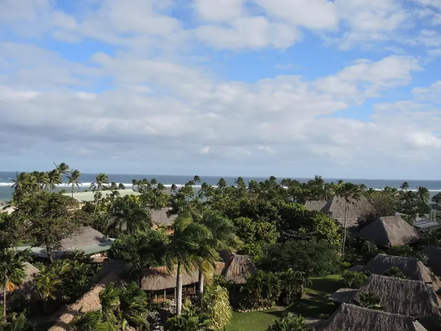 The image shows a view of the ocean from the top of a hill, with a number of trees, huts, and grass...