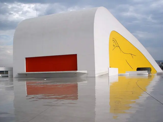 The image shows a large white building with a yellow and red mural on the side of it, surrounded by...