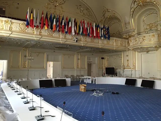 The image shows a large room with a long table and chairs in it. On the table there are microphones...