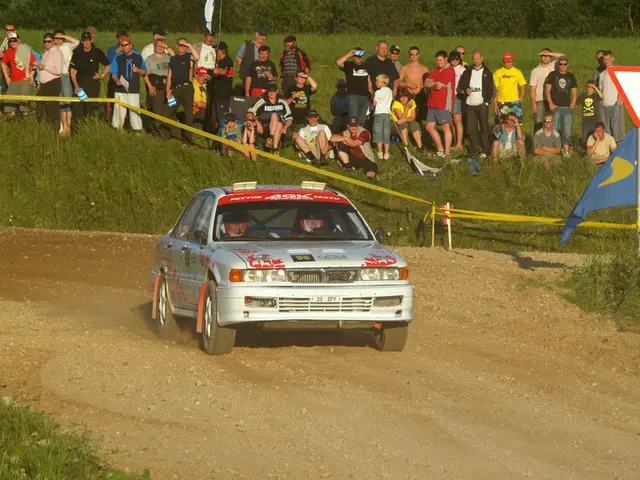 The image shows a rally car driving down a dirt road in front of a crowd of people, some of whom...