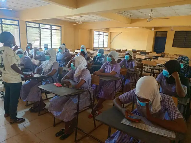 The image shows a classroom full of students wearing face masks, sitting on benches and some...