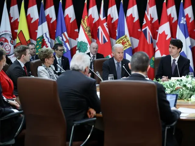 The image shows a group of people sitting around a table with flags in the background. On the table...