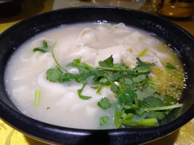 The image shows a bowl of wonton soup with coriander leaves on top, sitting on a table with a cloth...