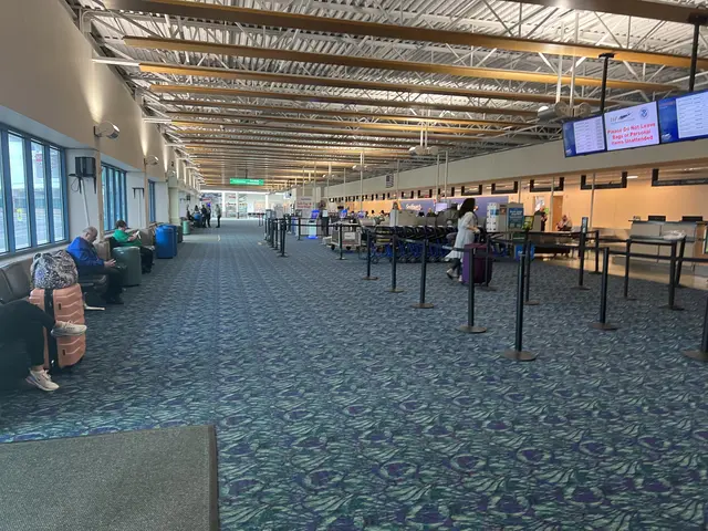 The image shows a long line of people waiting in an airport terminal, with people sitting on chairs...