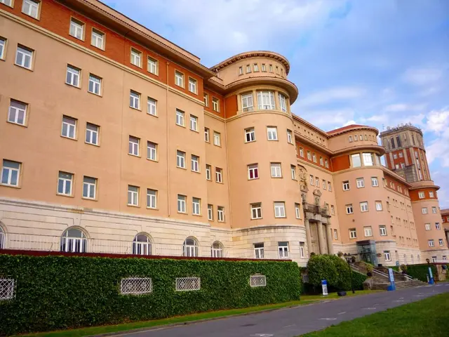 The image shows a large building with many windows on the side of it, surrounded by a road, grass,...