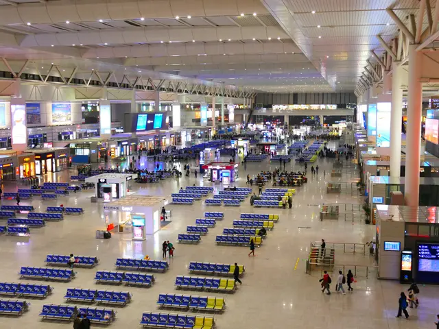 The image shows an inside view of an airport terminal with many people walking around, blue chairs,...
