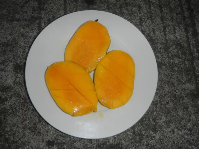 The image shows three mangoes on a white plate placed on a table. The mangoes are cut in half,...